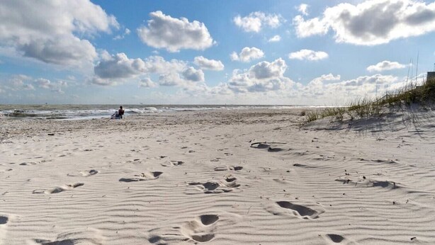 Een zandstrand met voetafdrukken, een zittend persoon en een heldere lucht met wolken.