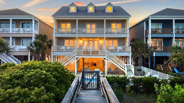 Un hotel de varios pisos frente a la playa, con balcones, piscina y un paseo de madera que conduce a la entrada.