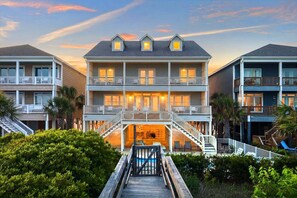 Un hotel de varios pisos frente a la playa, con balcones, piscina y un paseo de madera que conduce a la entrada.