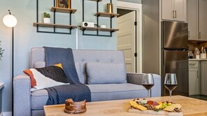 A modern living room with a grey sofa, a wooden coffee table with a cheese board, and a kitchen area with a stainless steel refrigerator. - Cozy 1 BR ADU | Private Access | Projector Theater (Windham)