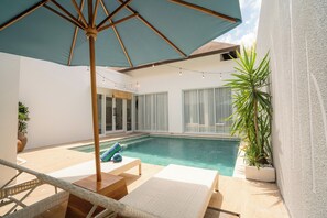 Un área junto a la piscina con sillones de descanso, una sombrilla grande y una piscina pequeña.