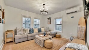 A modern living room with a grey sofa, patterned cushions, a coffee table, and a large window. - Tiki House  - Barefoot Breeze Walk to Beach (Treasure Island)