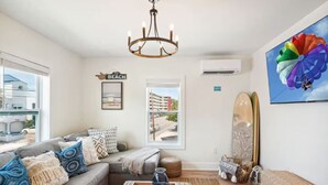 A living room with a grey sofa, a chandelier, a beach-themed sign, a surfboard, and a colorful kite painting. - Tiki House  - Sandy Toes Escape Walk to Beach (Treasure Island)