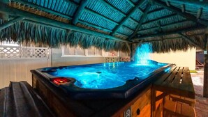 A hot tub under a thatched roof, surrounded by wooden furniture and a white fence. - Tiki House  - Sandy Toes Escape Walk to Beach (Treasure Island)