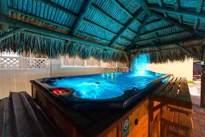 Un jacuzzi bajo un techo de paja, rodeado de mobiliario de madera y una cerca blanca.