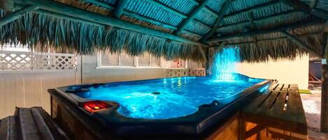 Un jacuzzi bajo un techo de paja, con una terraza de madera y una valla blanca al fondo.
