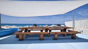 Une table de pique-nique en bois, avec des bancs, est installée sur une surface en béton, offrant une vue sur une plage rocheuse et sur une clôture en bois en arrière-plan.