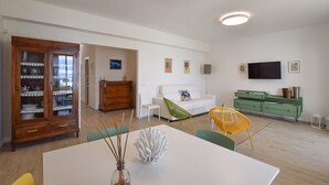 Un salon moderne avec un canapé blanc, des fauteuils verts et jaunes, un buffet en bois contenant de la vaisselle, et un téléviseur à écran plat fixé au mur.