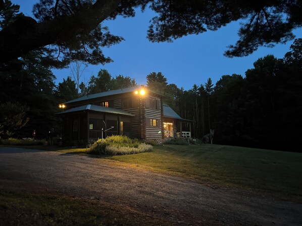 Une cabane en rondins surmontée d’un toit en métal, entourée d’arbres et bordée d’une pelouse bien entretenue.