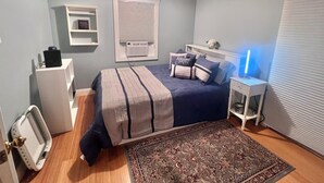 A bedroom with a bed, a bedside table, a nightstand, a radiator, a shelf, and a window with blinds.