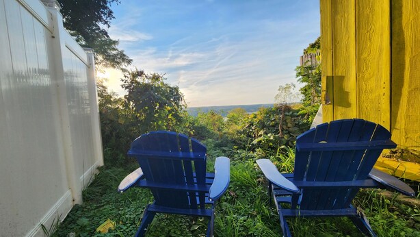 Two blue Adirondack chairs facing a lush garden with a view of distant hills. - The Seneca Sunsets Studio (Burdett)