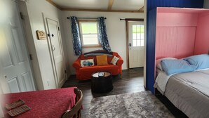 A room with a bed, a red sofa, a small table, and a window with curtains. - The Seneca Sunsets Studio (Burdett)