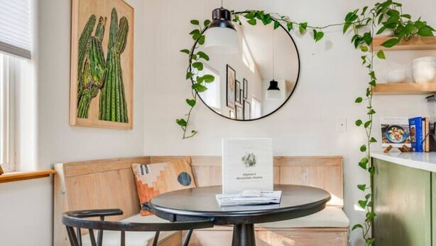 Une pièce chaleureuse avec un miroir rond, un banc en bois, une petite table ronde et un tableau représentant un cactus accroché au mur.