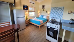 A compact room with a bed, a small table, a chair, a refrigerator, an oven, and a ceiling fan. - Sunny studio in Esperanza (Vieques)