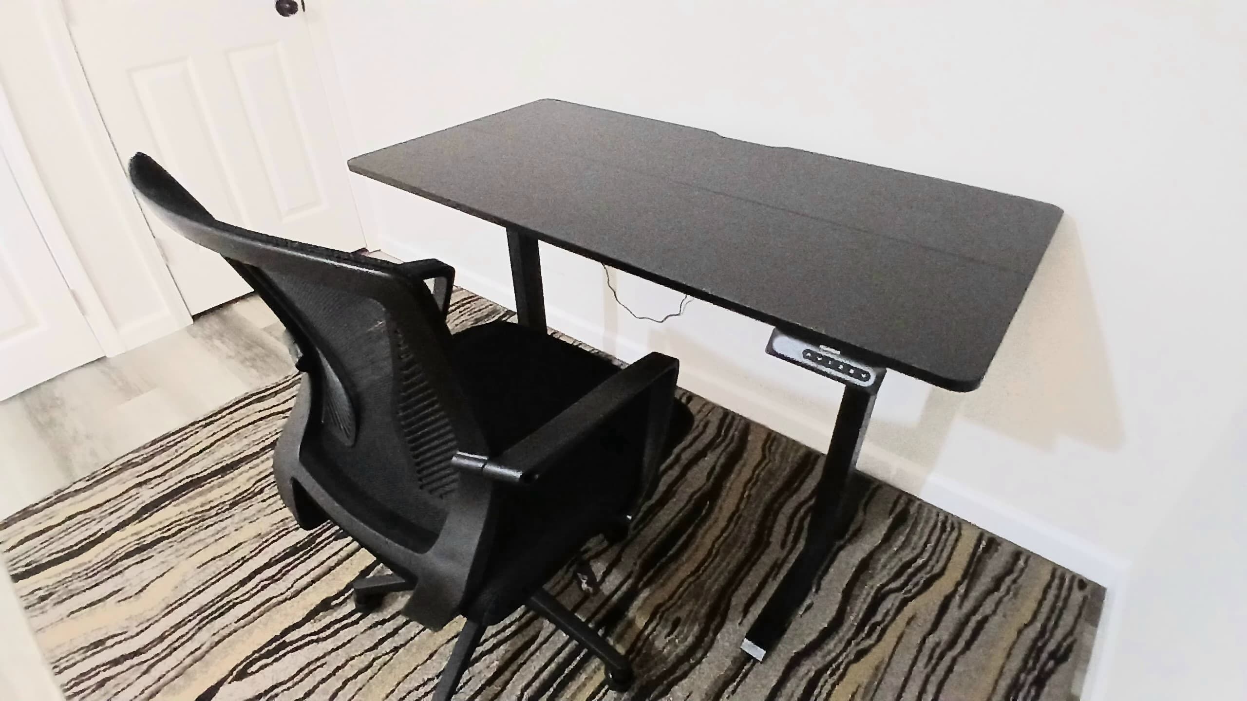 Un petit bureau de couleur sombre, accompagné d’une chaise, est placé contre un mur blanc, devant un tapis à motifs.