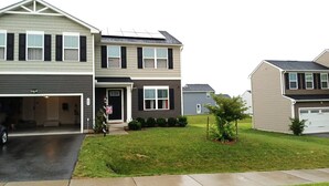A two-story residential house with a garage, a small front yard with grass and a tree, and solar panels on the roof.