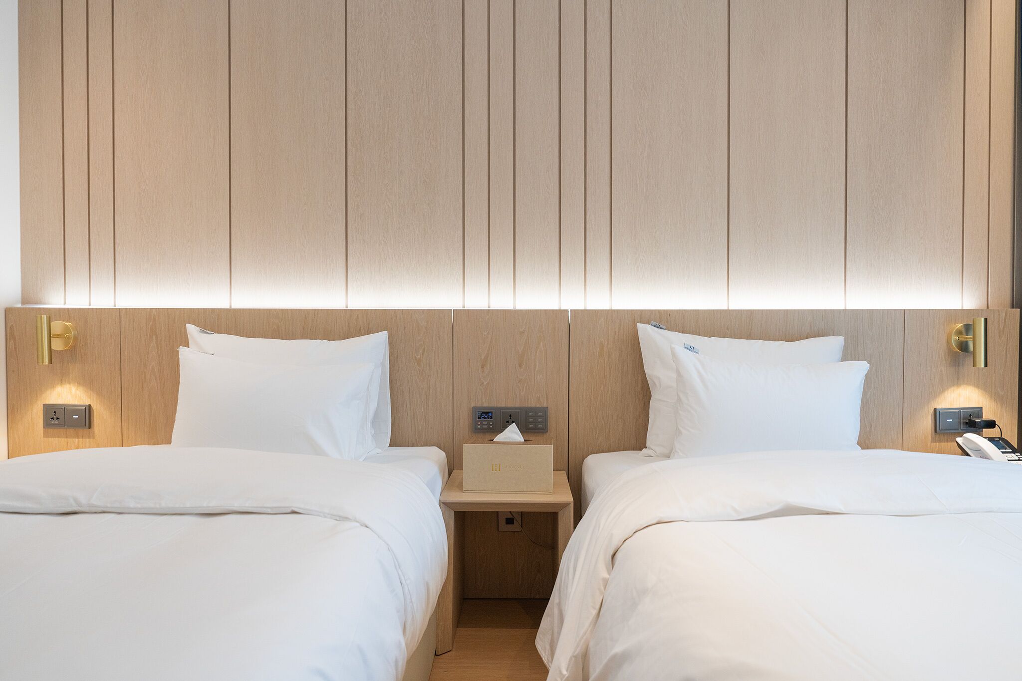 Une chambre d’hôtel avec deux lits, une table de chevet avec une lampe, un téléphone et un panneau de commande au mur.