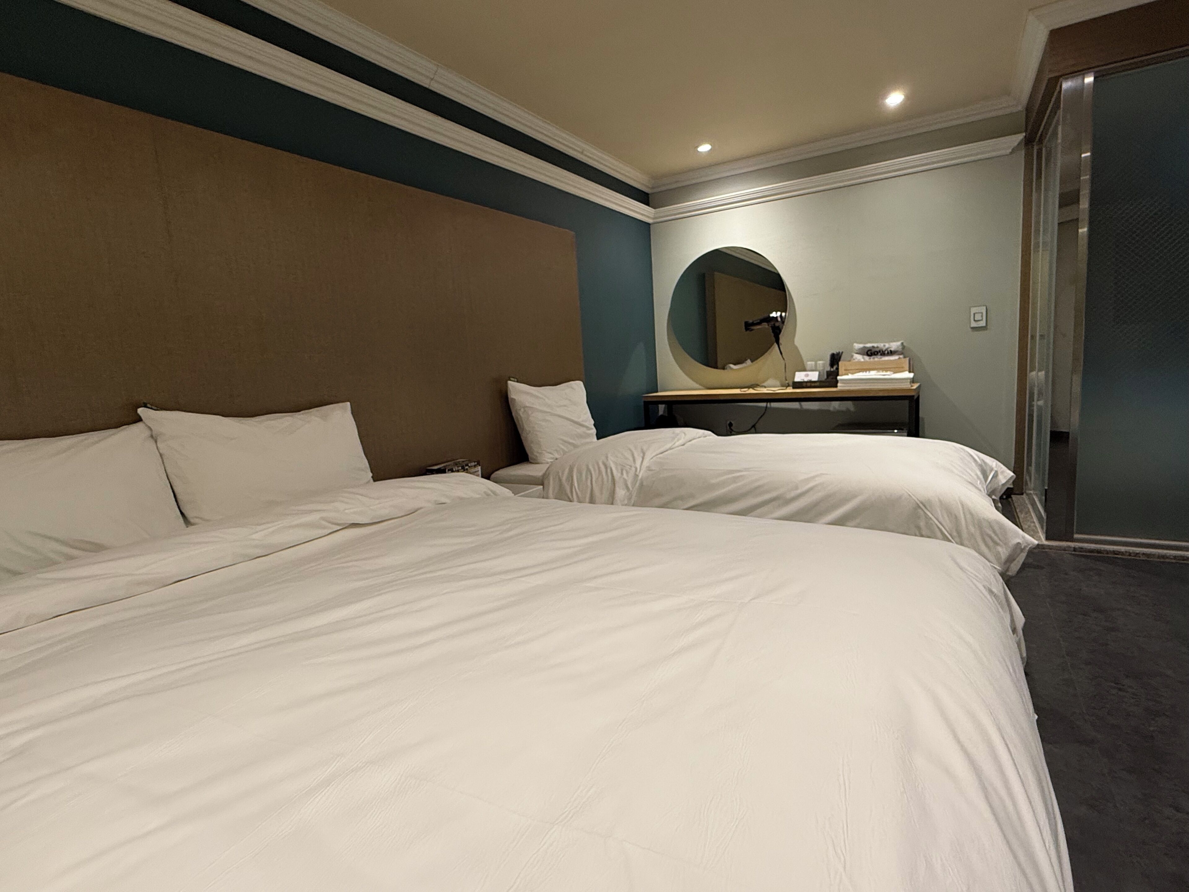 Habitación de hotel con dos camas, un espejo redondo, una mesita con lámpara y una puerta corredera.
