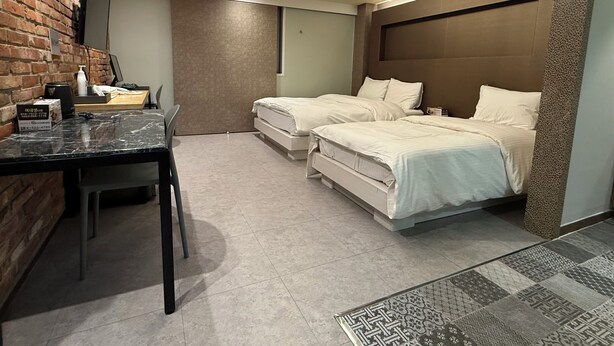 A hotel room with a brick wall, a desk with a chair, two beds, and a patterned rug.