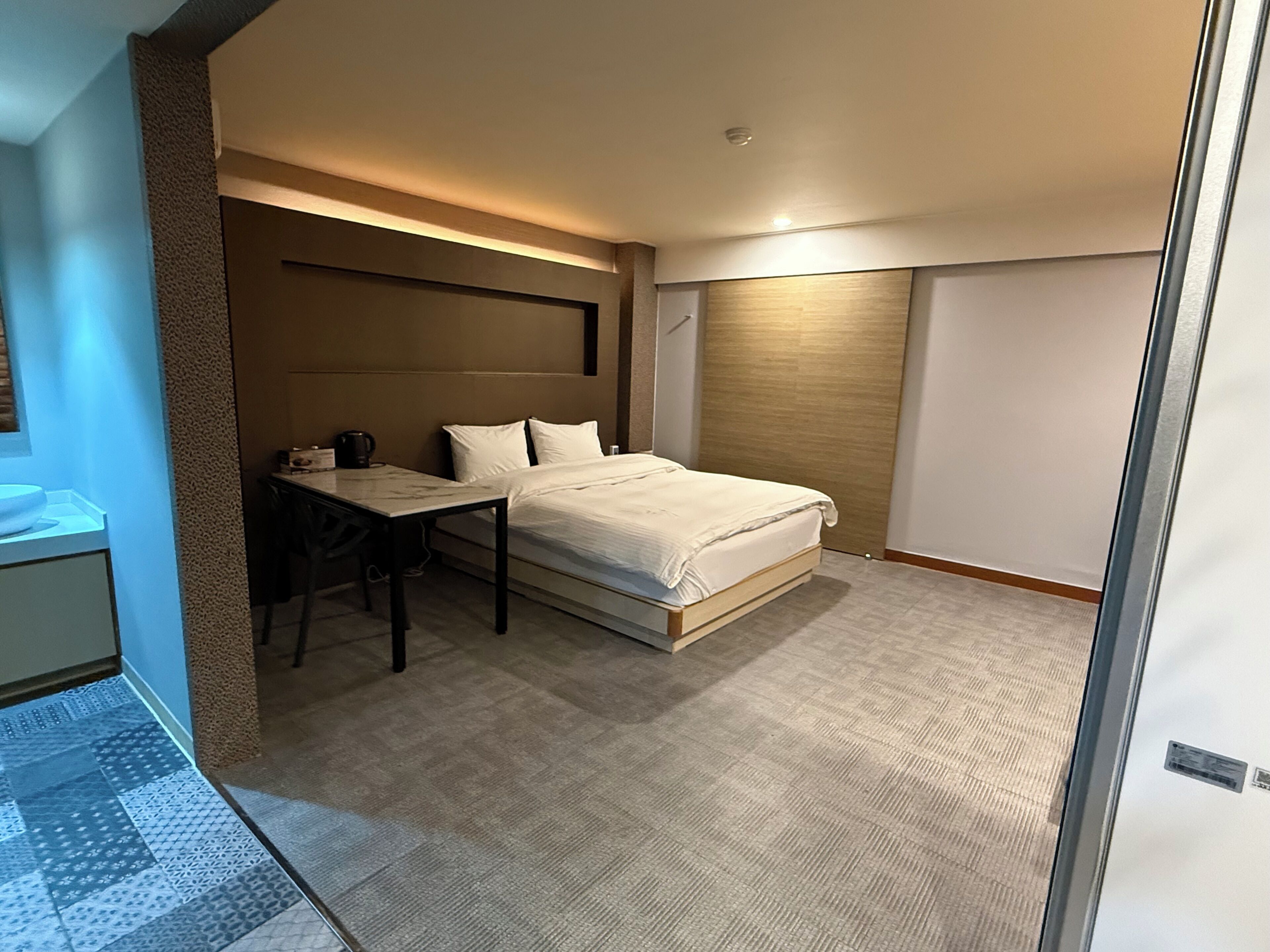 A hotel room with a bed, a desk, a chair, a small table, and a bathroom area.