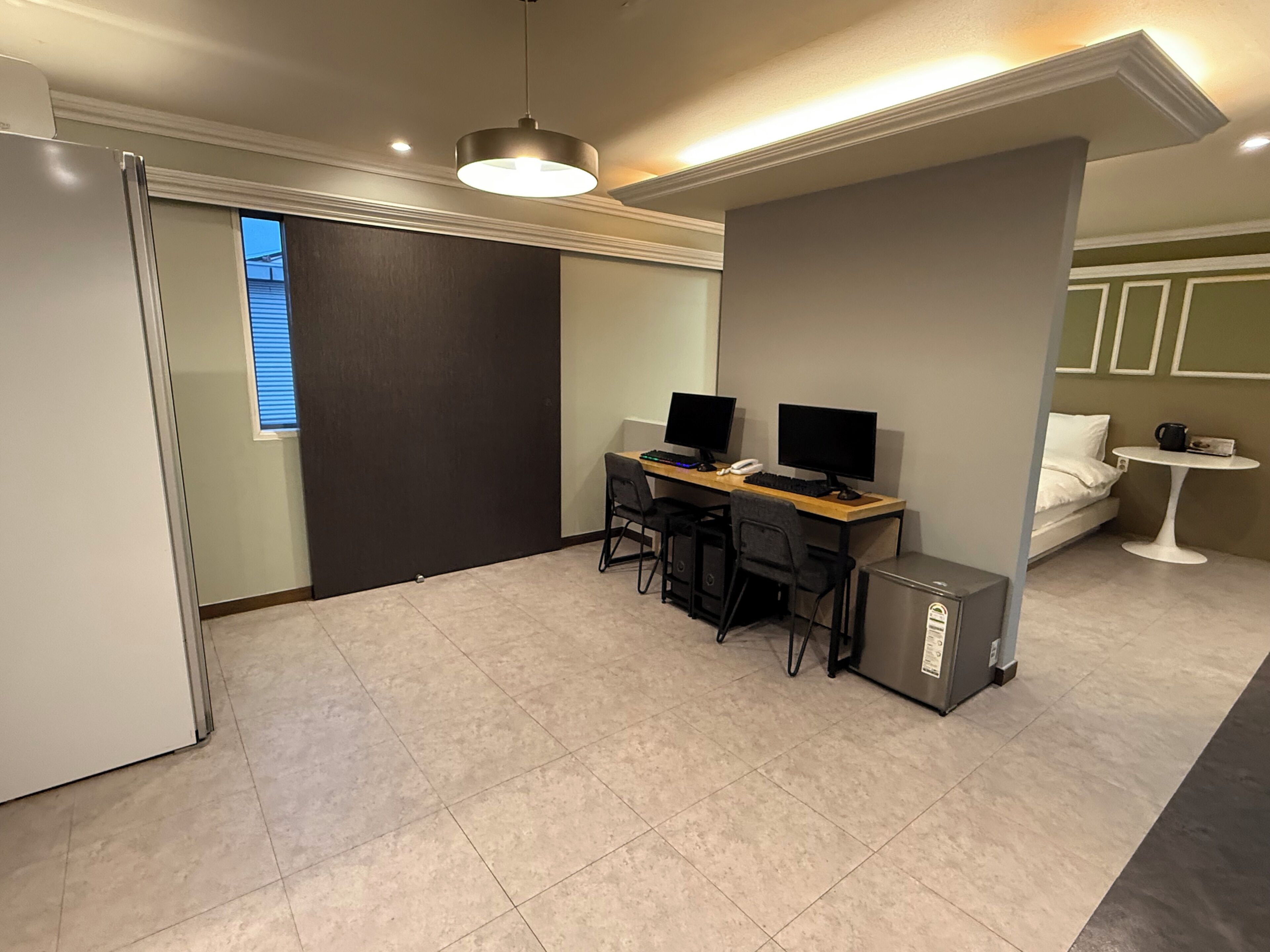 Habitació petita amb escriptori, dos monitors, nevera i llit.