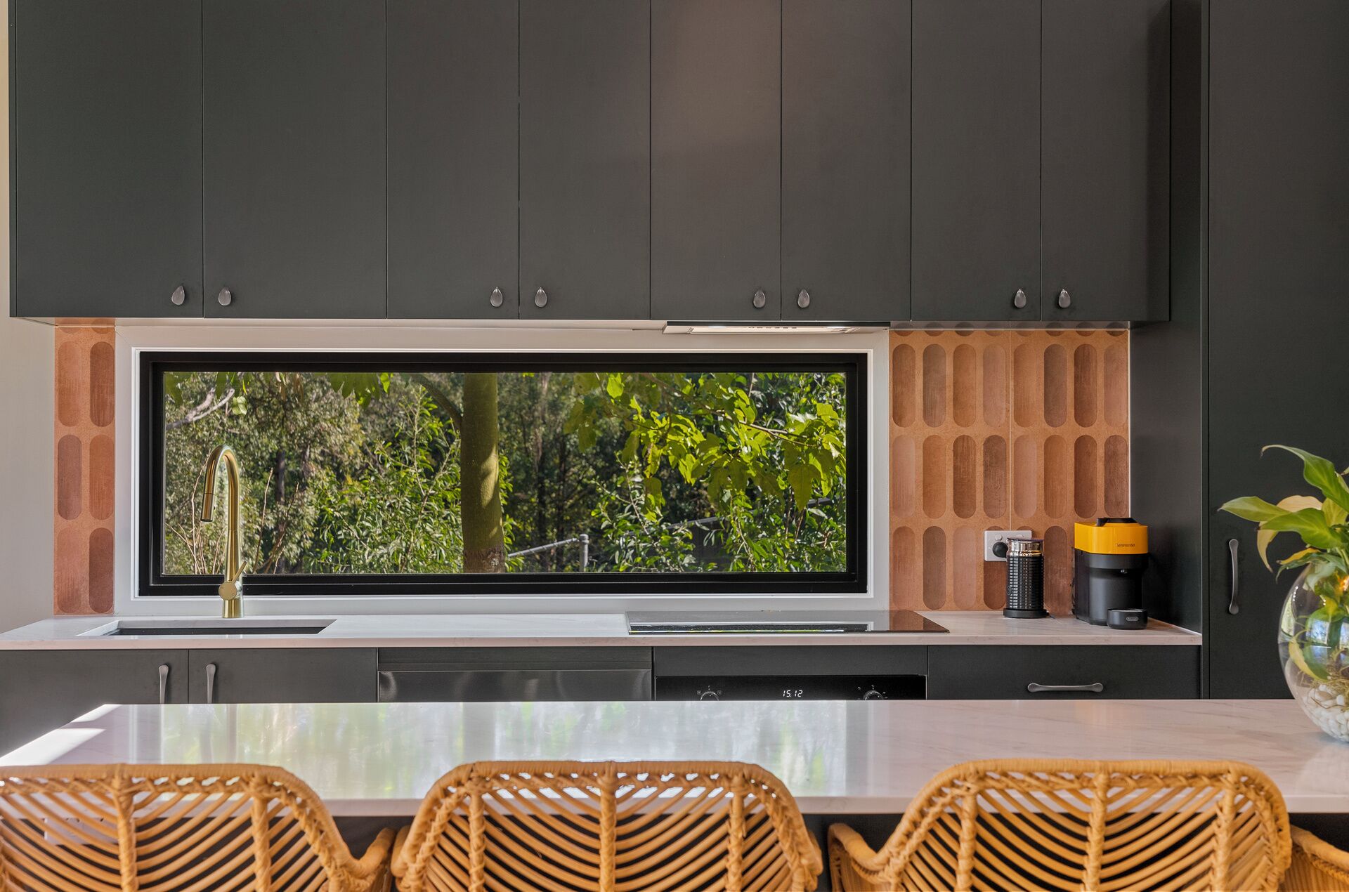 Una cocina moderna con armarios oscuros, un ventanal con vista a la vegetación, una cafetera y un florero con plantas.