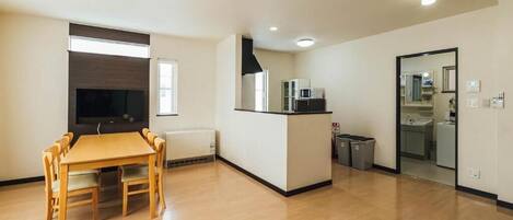 Un salón amplio con una mesa y sillas de madera, un televisor empotrado y una zona de cocina con microondas y botes de basura.