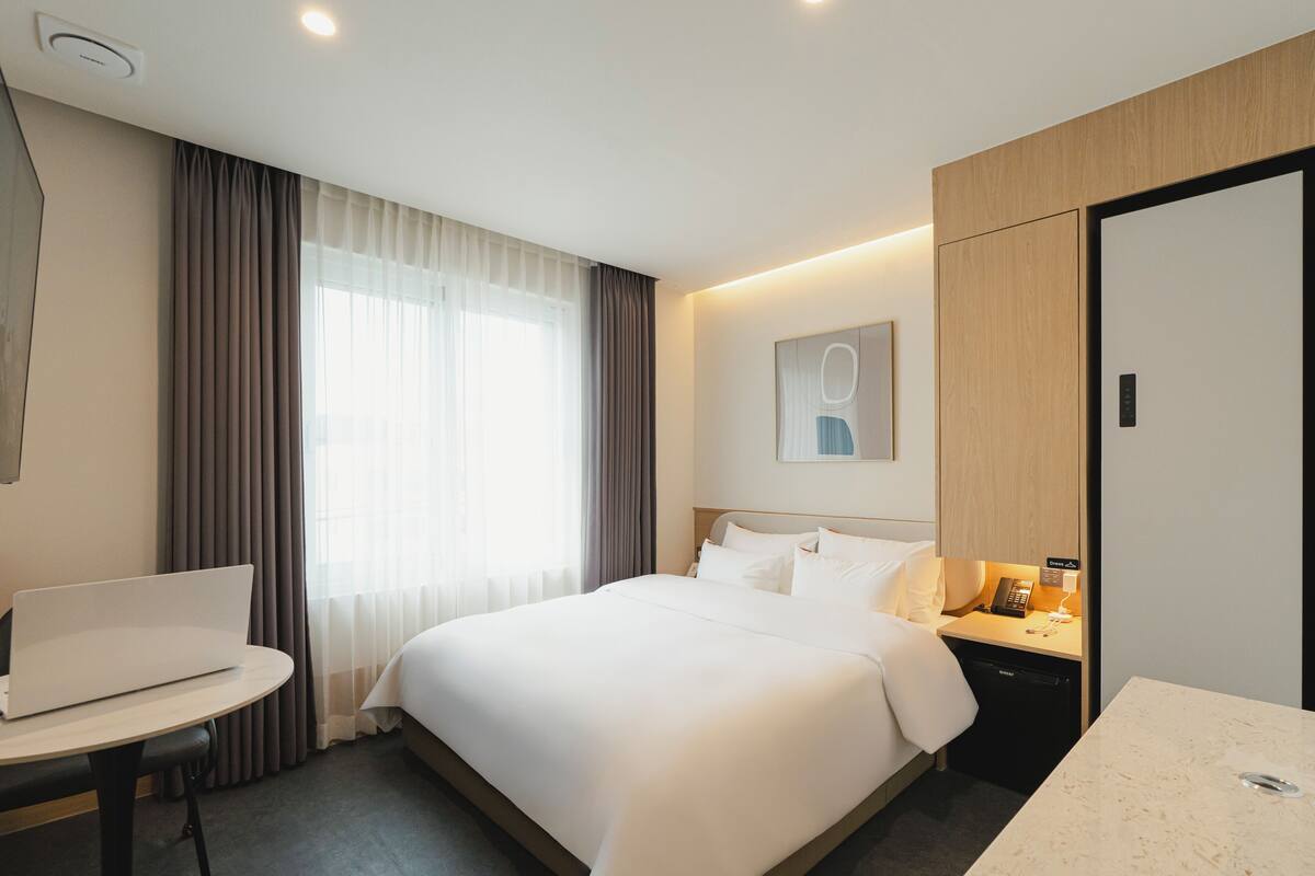 Habitación de hotel con cama, escritorio con una computadora portátil, mesita de noche con teléfono y ventana grande con cortinas.