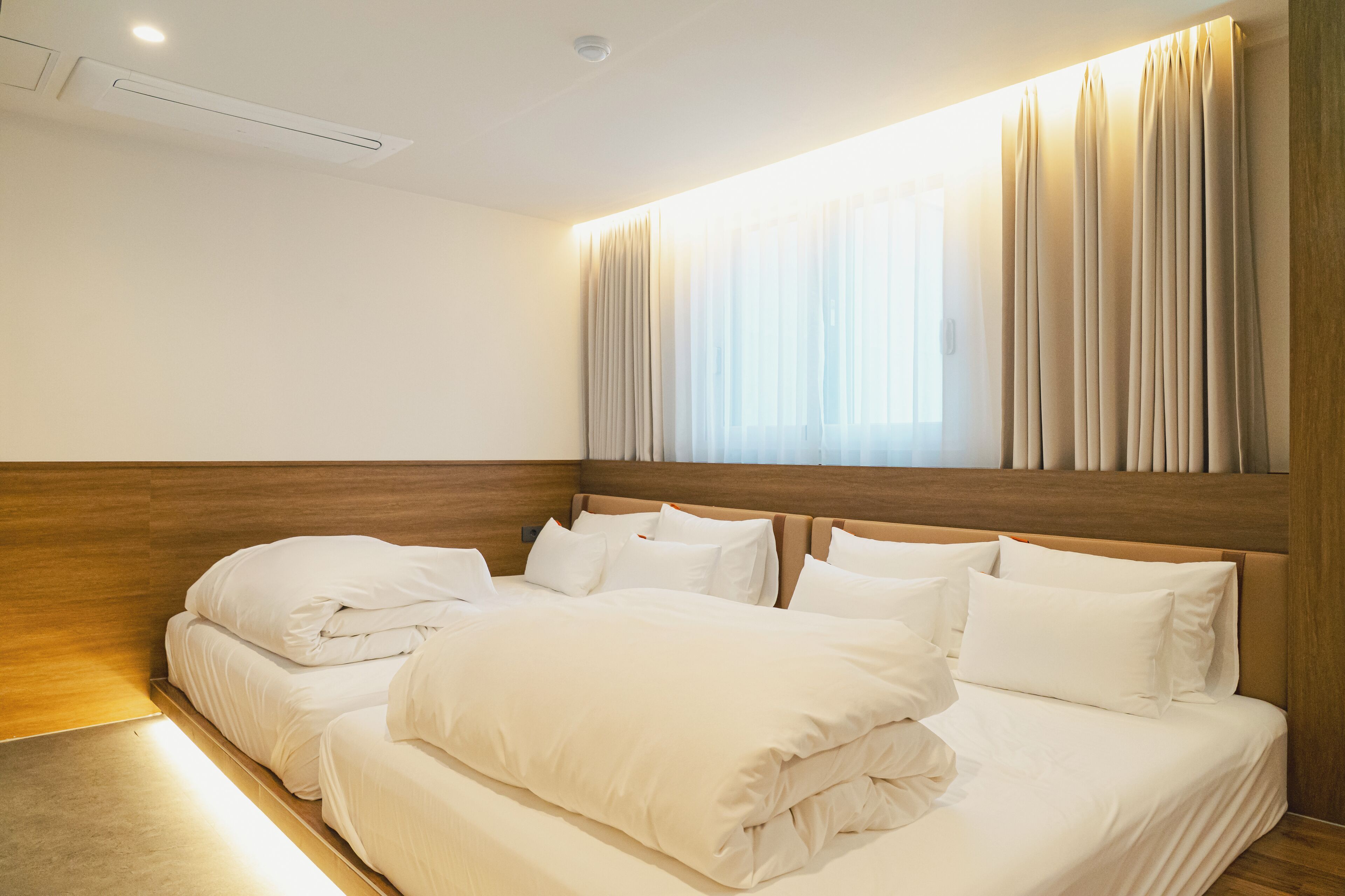 Een hotelkamer met een houten hoofdbord, een groot bed met wit beddengoed, en een raam met dunne gordijnen.