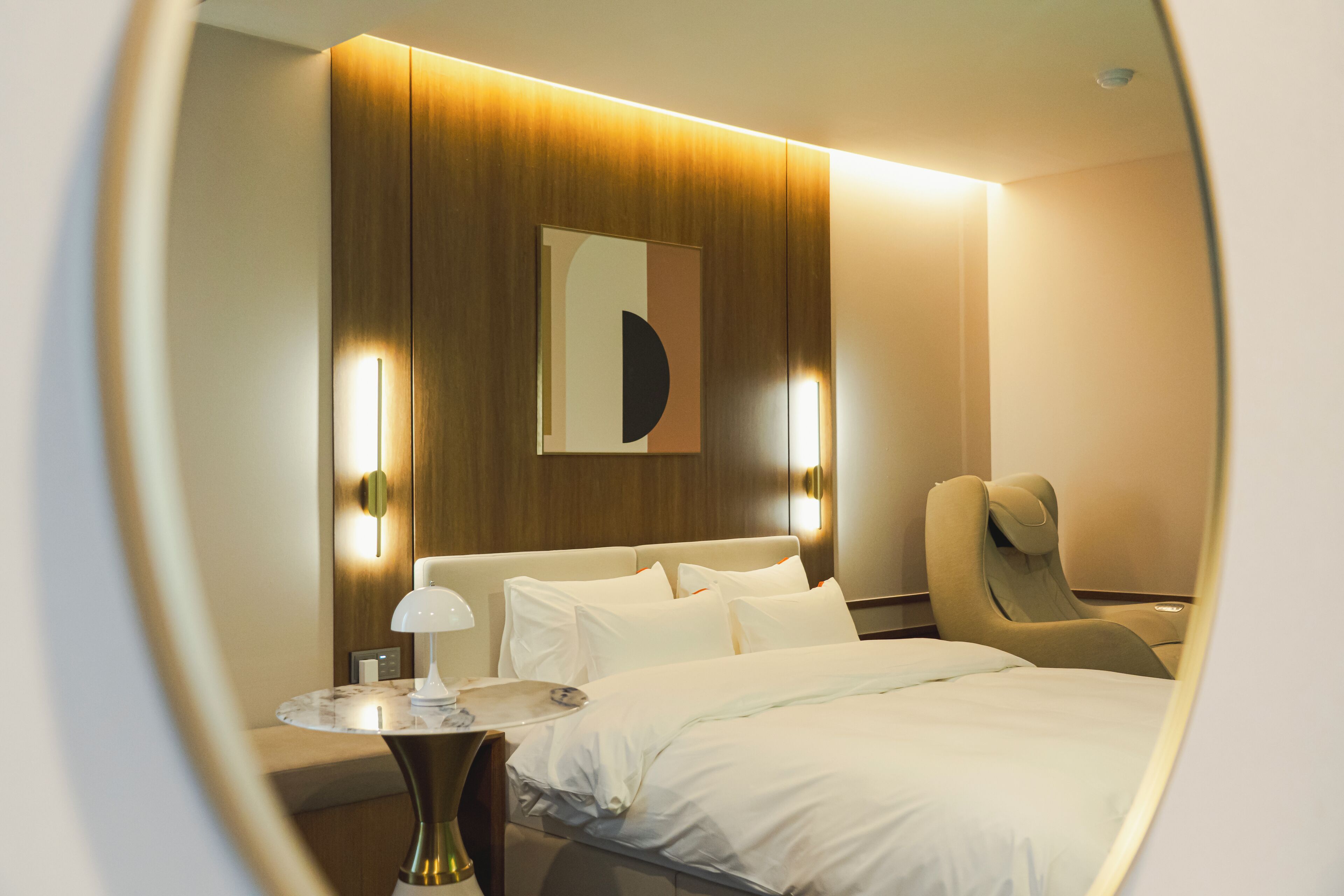 Quarto de hotel com cama, uma cadeira, uma mesinha e luminária fixada na parede.
