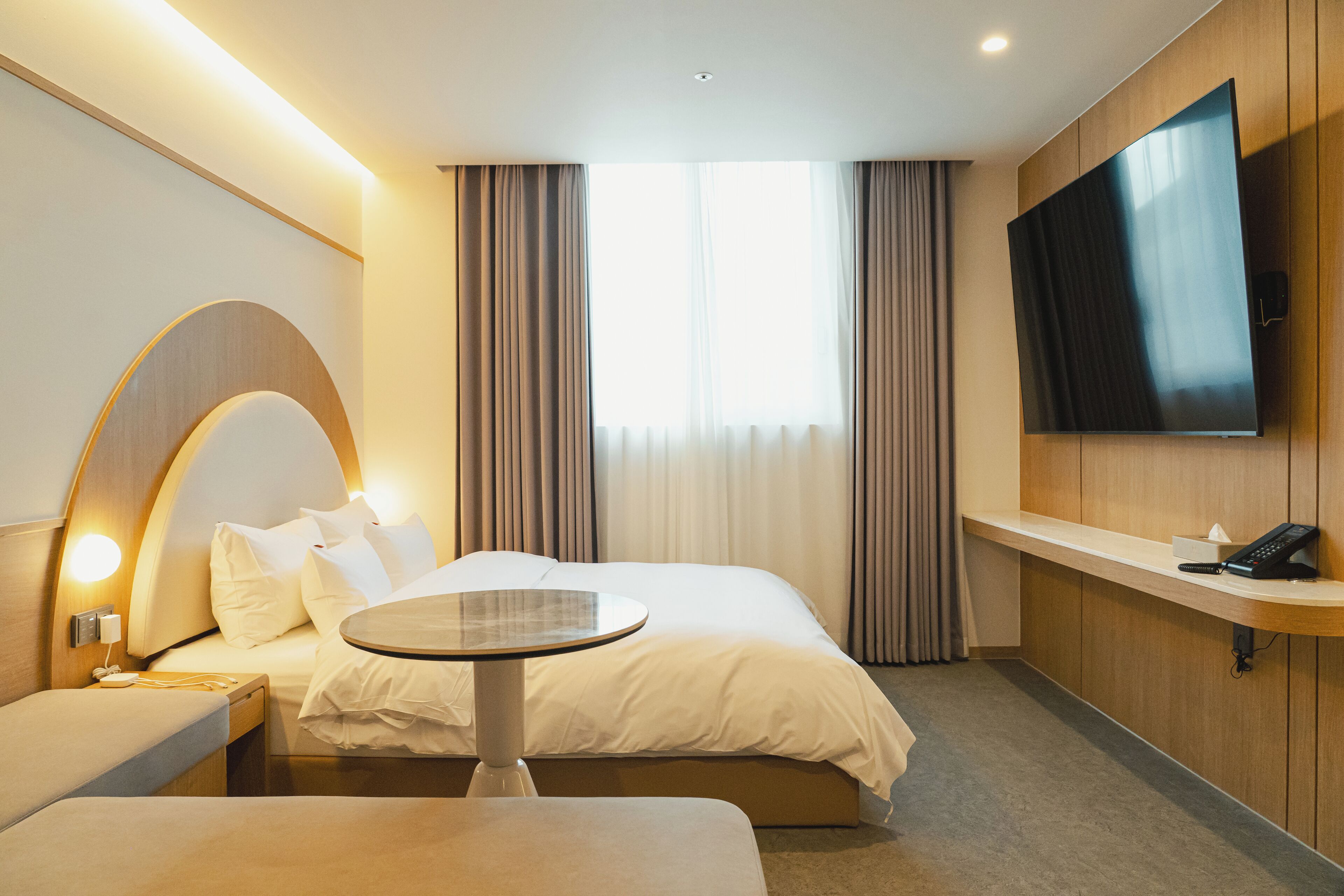 A modern hotel room with a large bed, a bedside table, a round table, a TV mounted on the wall, and a window with curtains.