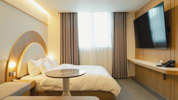 A modern hotel room with a large bed, a bedside table, a round table, a TV mounted on the wall, and a window with curtains.