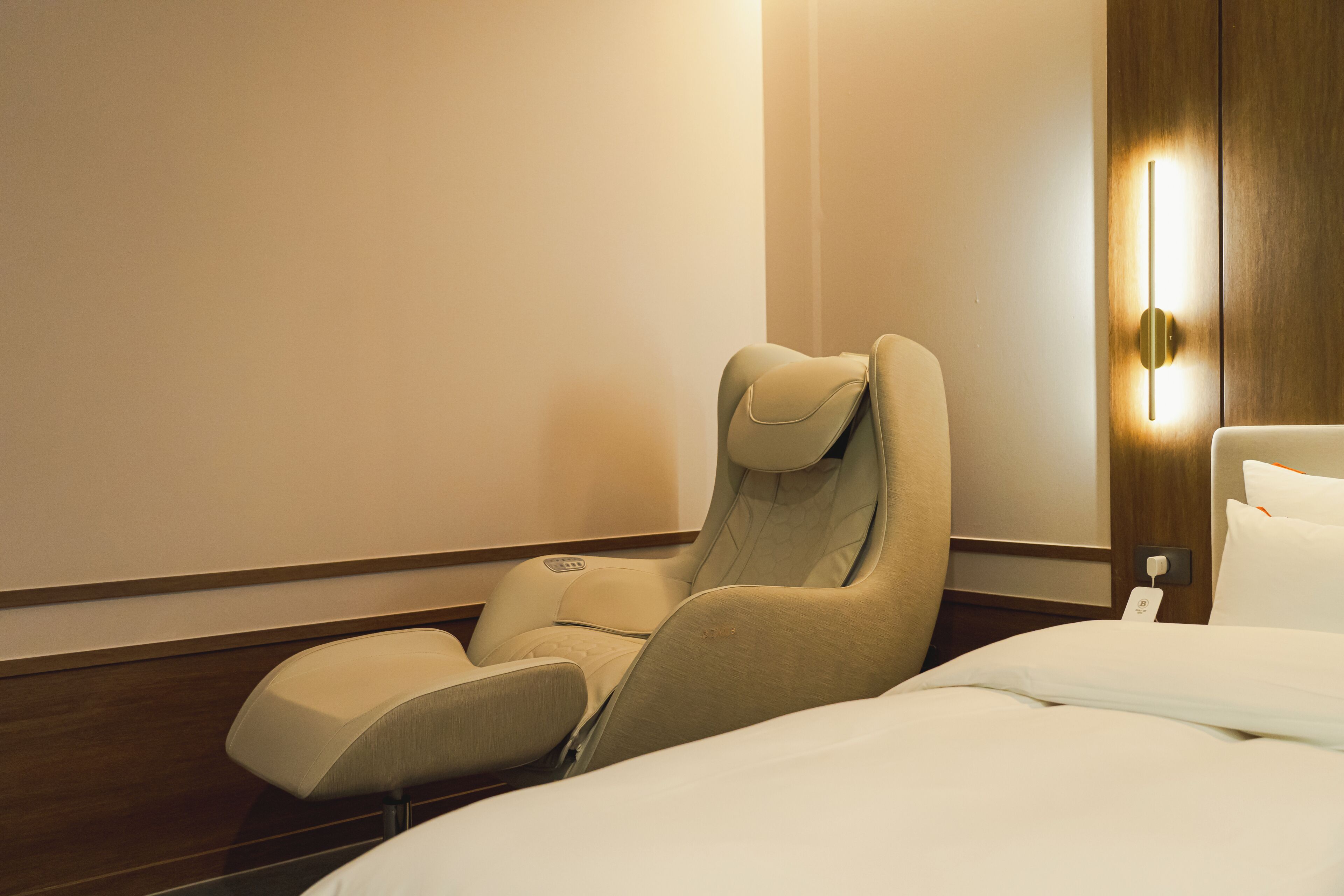 A hotel room with a massage chair, a bed, and a wall-mounted light fixture.