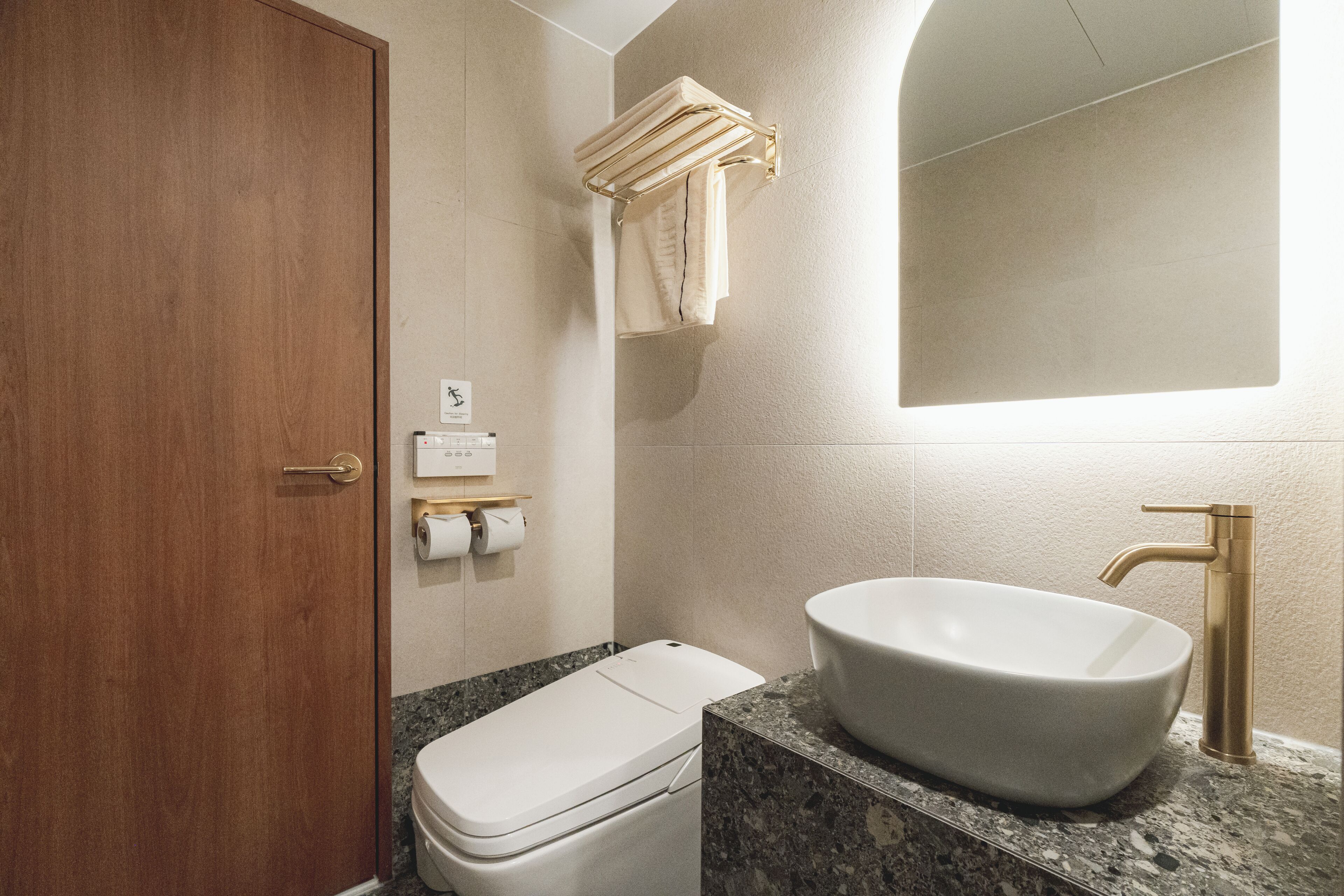 Un baño con puerta de madera, inodoro blanco, lavamanos con grifo dorado y toallero con toallas.