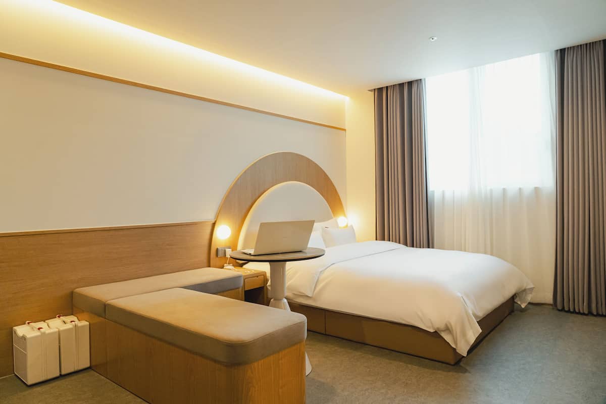 Habitación de hotel con cama, escritorio con computadora portátil, zona de estar y ventana grande con cortinas.