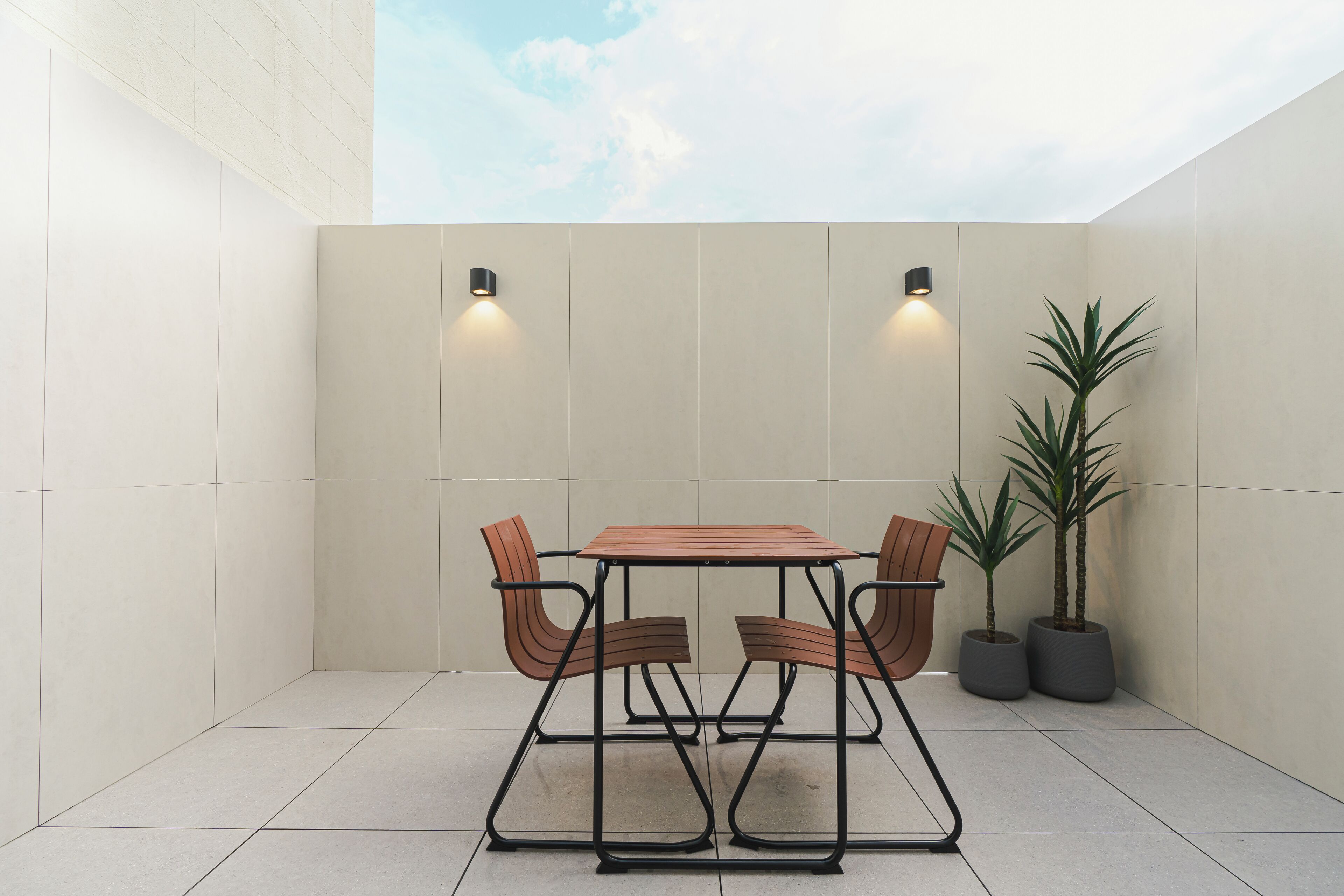 Un pequeño patio exterior con una mesa y sillas, dos luces fijadas a la pared y dos macetas con plantas.
