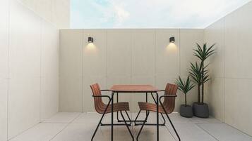 Un pequeño patio exterior con una mesa y sillas, dos luces fijadas a la pared y dos macetas con plantas.