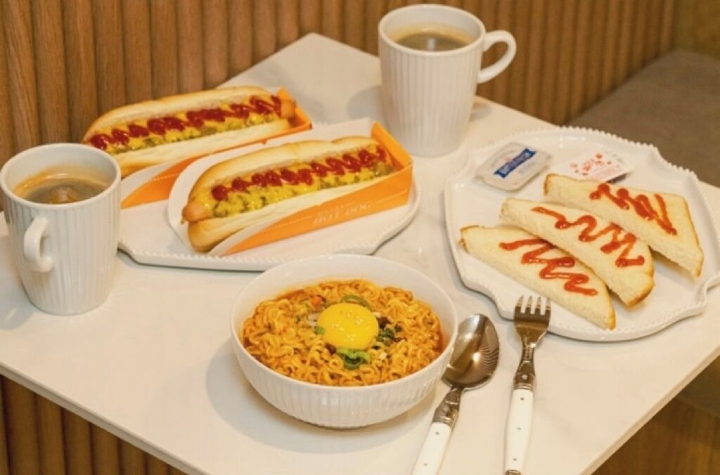A table with two hot dogs, a bowl of noodles, toast with ketchup, and two cups of coffee.