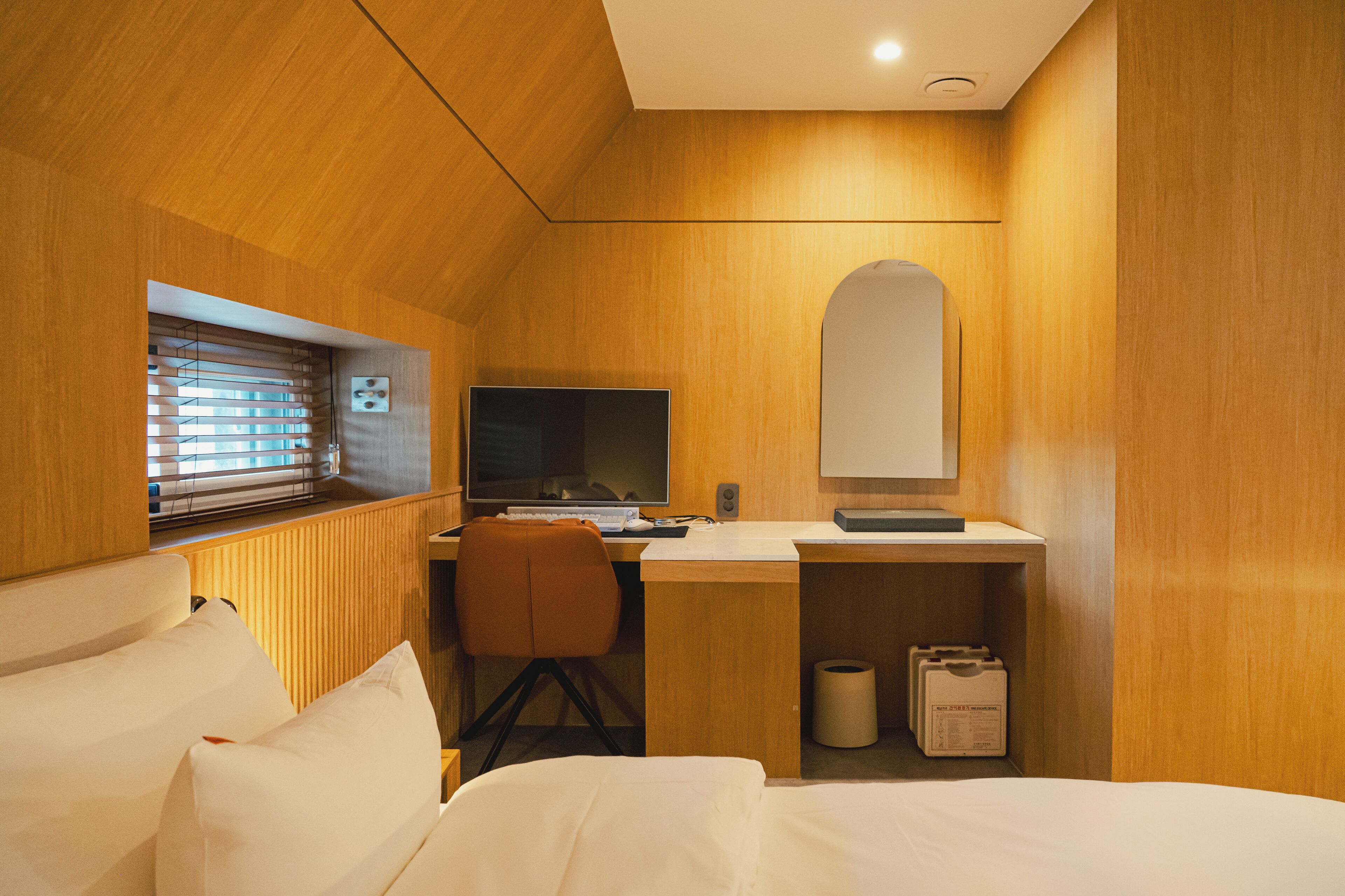 Quarto de hotel com decoração em madeira, uma cama, uma escrivaninha com computador, uma cadeira e uma janela com persianas.