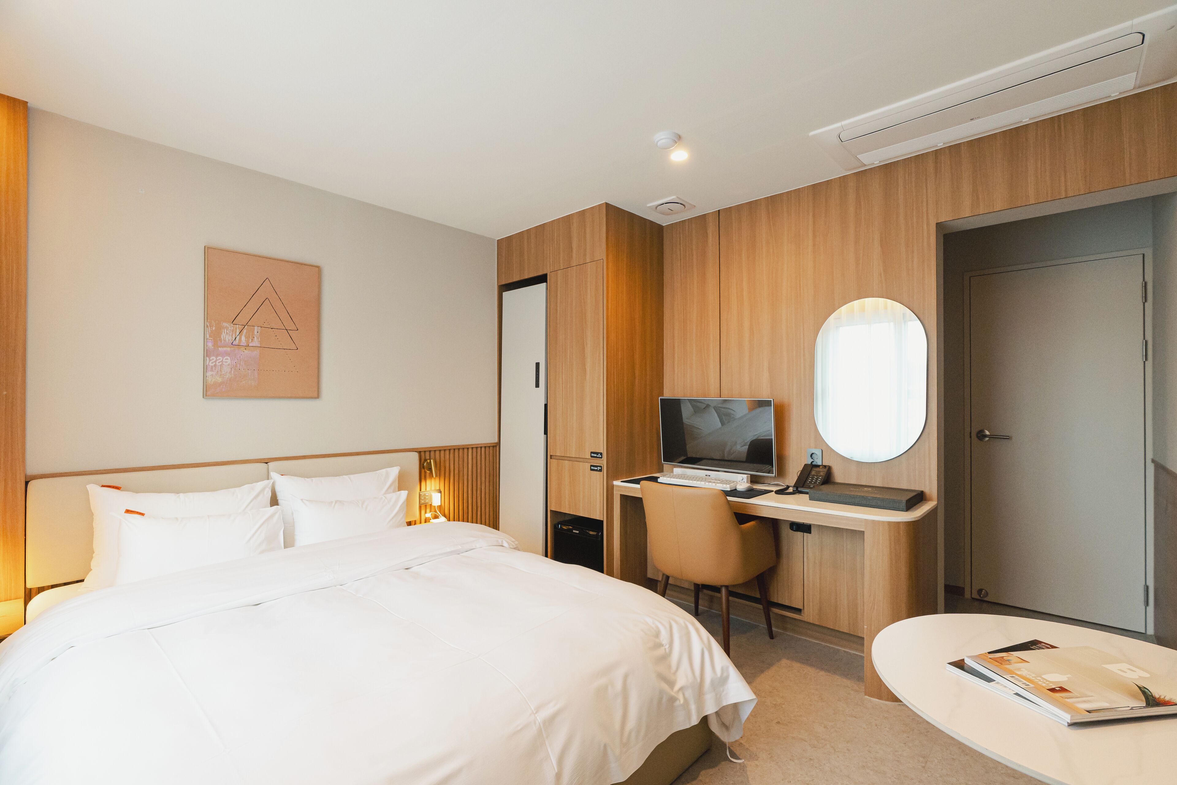 A hotel room with a bed, a desk with a computer, a chair, a desk lamp, a mirror, and a door.