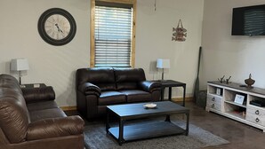 A living room with a brown leather couch, a black coffee table, a wooden-framed window, a ceiling fan, a wall clock, a TV, and a white cabinet with drawers.