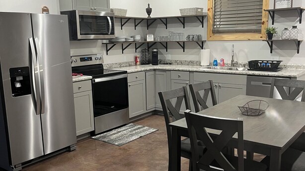 A modern kitchen with stainless steel appliances, a dining area with dark wooden chairs, and built-in shelves with kitchenware.