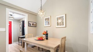 Un comedor con una mesa de madera, sillas y una araña. Hay cuadros en la pared y una puerta roja al fondo.