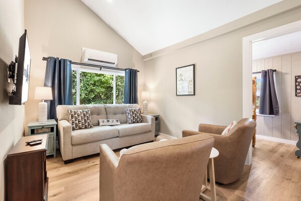 A living room with a sofa, two armchairs, a small table, and a TV. There is a window with curtains, an air conditioning unit above it, and a framed picture on the wall.