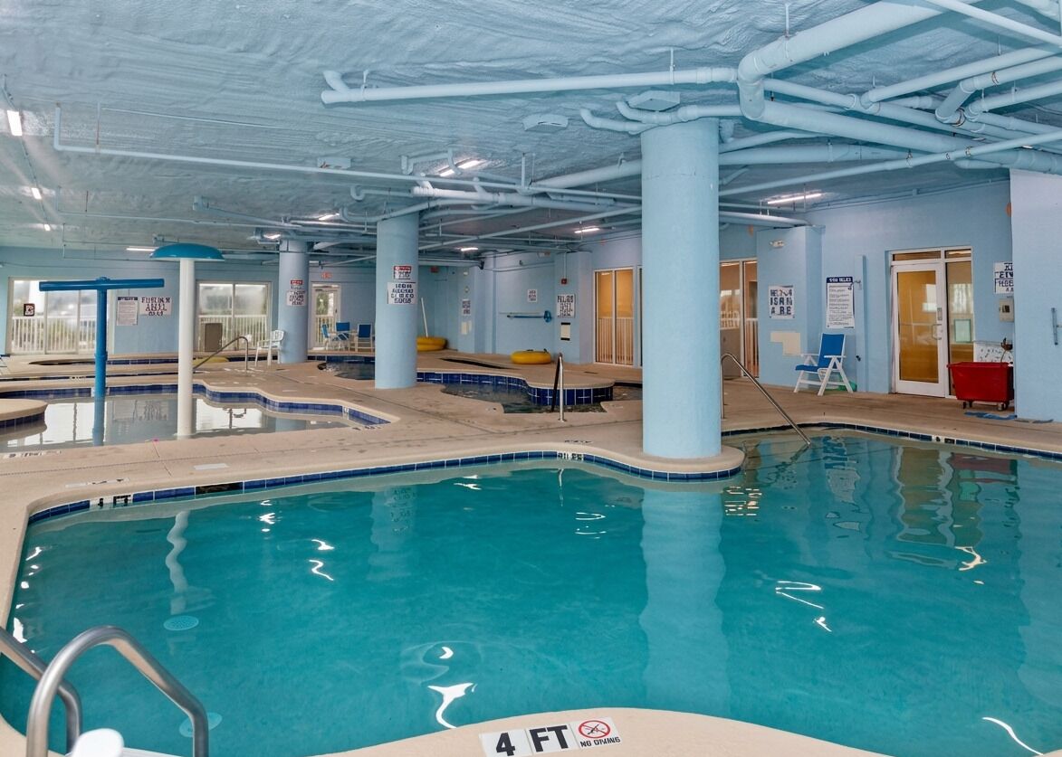Une piscine intérieure à l’eau claire, avec une indication de profondeur de 1,2 mètre et un plafond bleu où l’on peut voir les tuyaux.