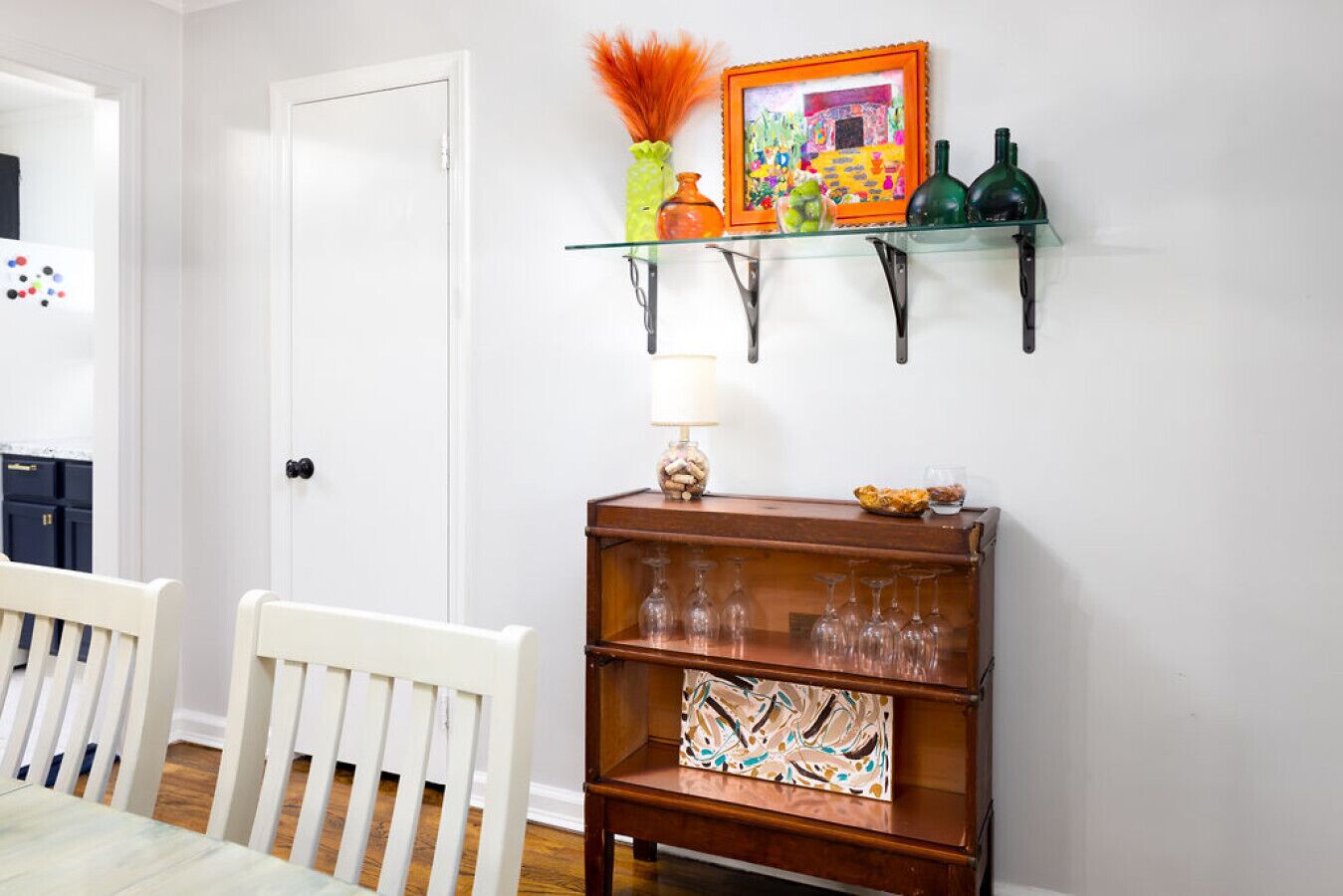 A dining area with a white chair, a wooden sideboard with glassware, and a wall-mounted shelf with decorative items.