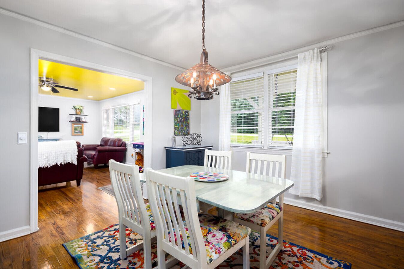 A dining area with a table set for four, surrounded by white chairs with colorful cushions. A chandelier hangs above the table, and a colorful rug is on the floor.