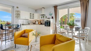 Una cocina moderna con armarios blancos, una zona de comedor con sillas amarillas y vistas a palmeras.