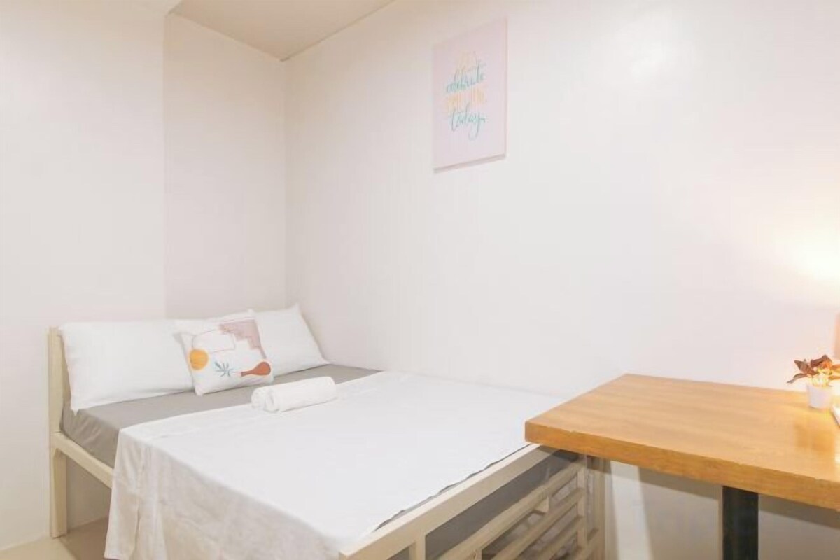 A small, clean room with a bed, a wooden table, and a wall-mounted light.