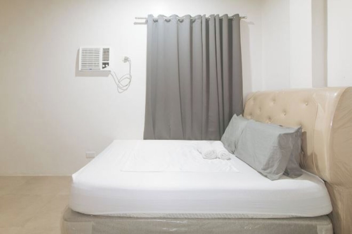 Una cama bien tendida con colchón y almohadas blancos, cabecera gris y un equipo de aire acondicionado instalado en la pared.